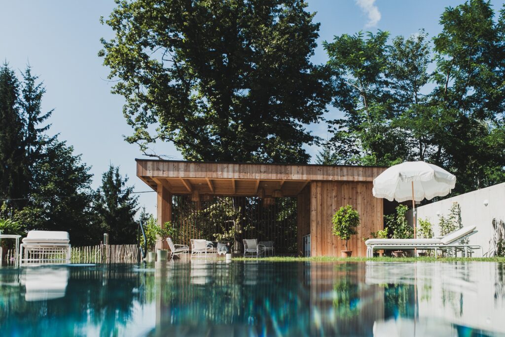 Außenpool mit spiegelndem Wasser, weiße Sonnenliegen und Schirm vor modernem Holzpavillon im grünen Hotelgarten unter hohen Bäumen – ruhige Wellness-Atmosphäre.