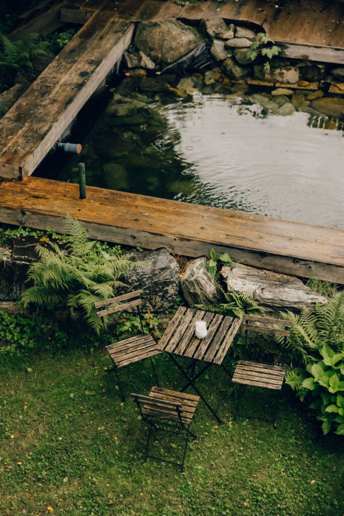 Rustikaler Gartenbereich im Waldhaus Rudolfshöhe mit Holztisch, Stühlen und Blick auf den Naturteich.