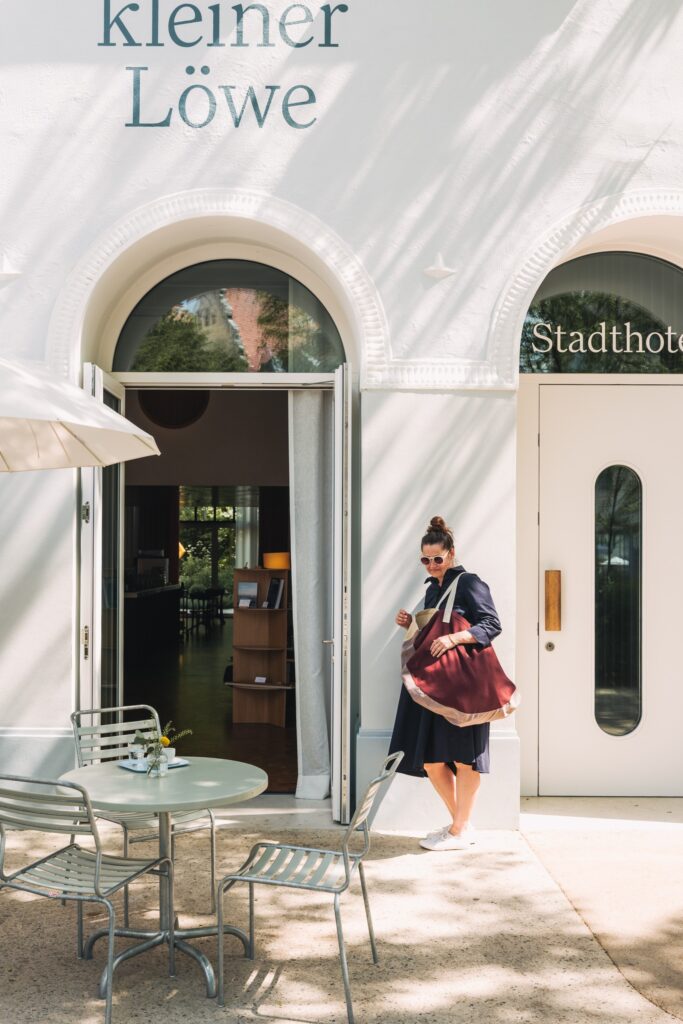 Eingang des Stadthotels kleiner Löwe in Bregenz mit Schriftzug und Rundbögen; ein Gast tritt mit der roten Badetasche aus dem Haus in die Sonne.